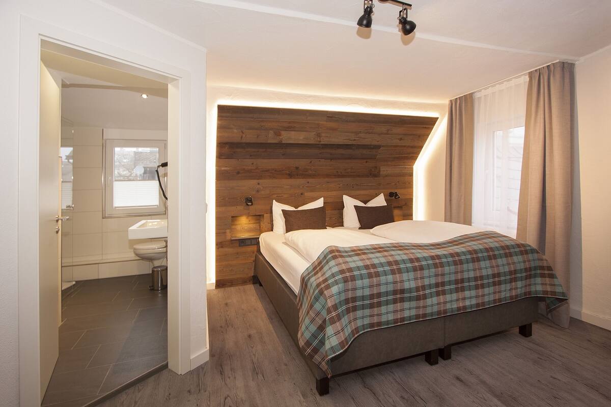 A bedroom with a wooden headboard, a bed with a plaid bedspread, a bedside table, and a window with curtains.