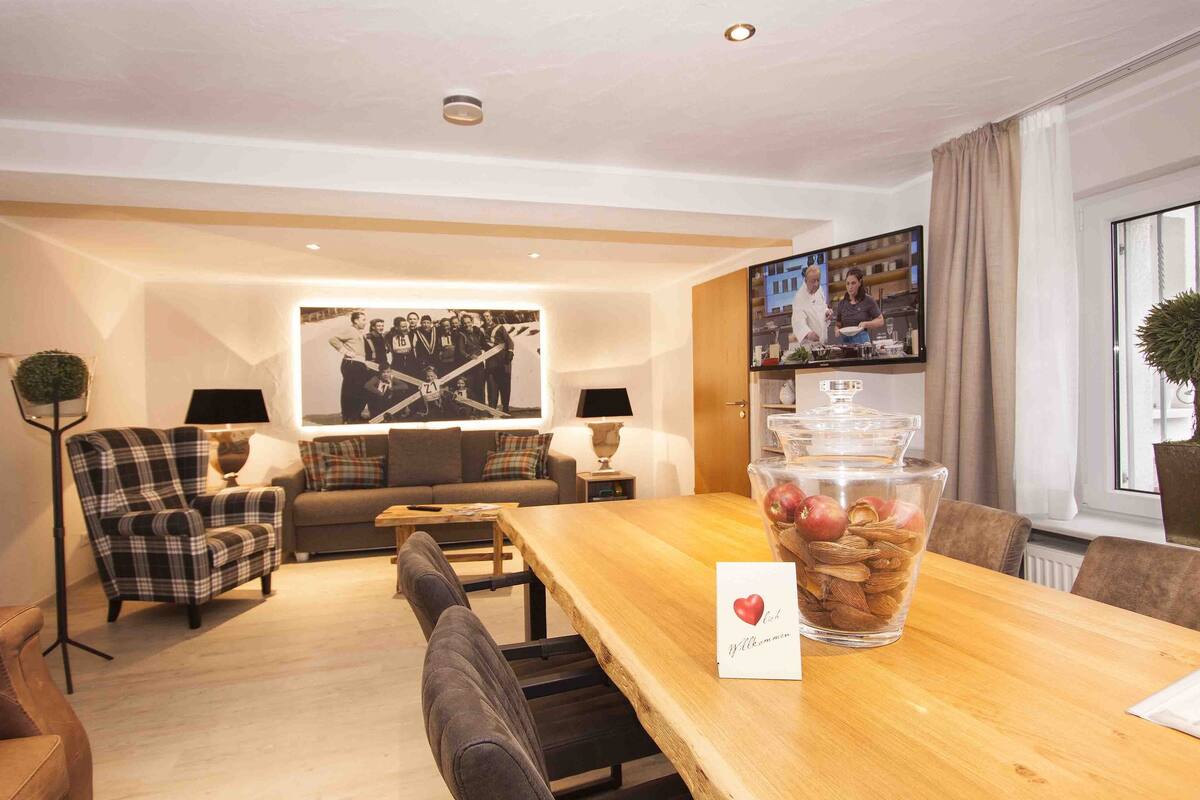 A modern hotel room with a wooden dining table, a plaid armchair, a sofa, and a large window with curtains.
