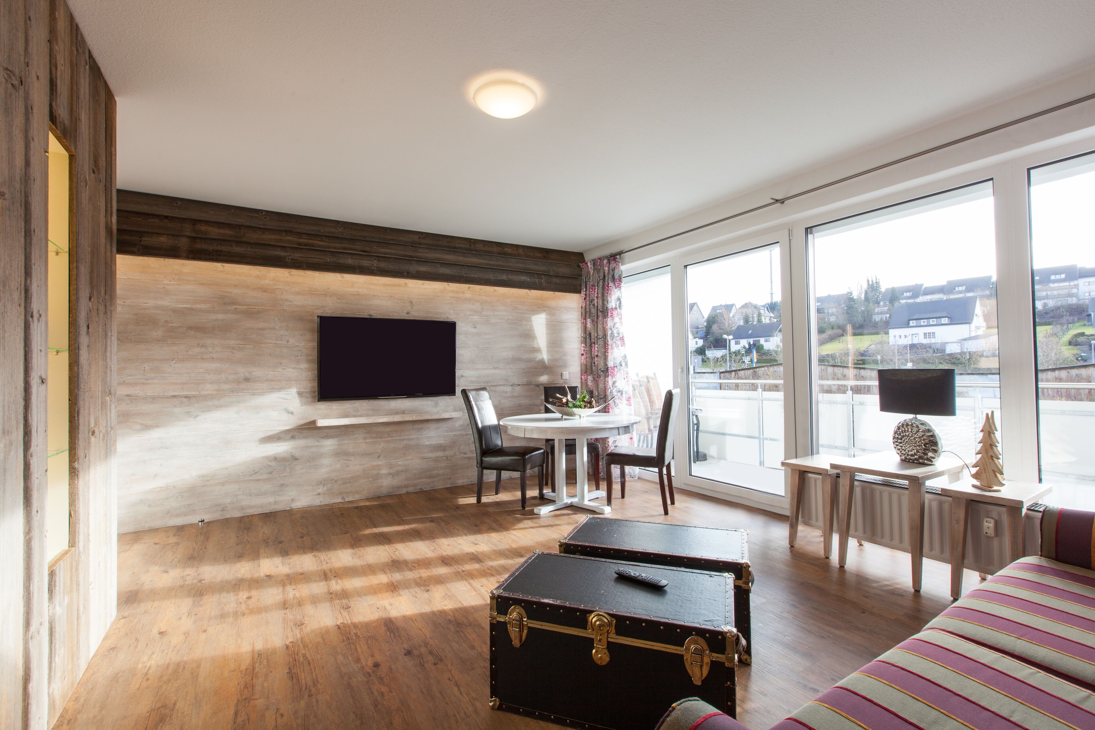 A modern room with a wooden floor, a flat-screen TV mounted on the wall, a dining table with chairs, a small desk with a lamp, and a large window offering a view of the city.