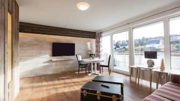 Une chambre moderne avec un plancher en bois, un téléviseur à écran plat fixé au mur, une table à manger avec des chaises, un petit bureau avec une lampe et une grande fenêtre donnant sur la ville.
