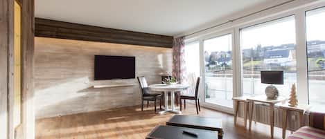 A modern room with a wooden floor, a flat-screen TV mounted on the wall, a dining table with chairs, a small desk with a lamp, and a large window offering a view of the city.
