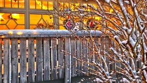 Una cerca de madera con ramas cubiertas de nieve frente a un edificio con ventanas iluminadas.