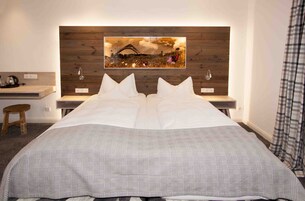 Una cama bien hecha con una colcha a cuadros, un cabecero de madera con una pintura de paisaje, una mesita de noche con una lámpara y un taburete pequeño.