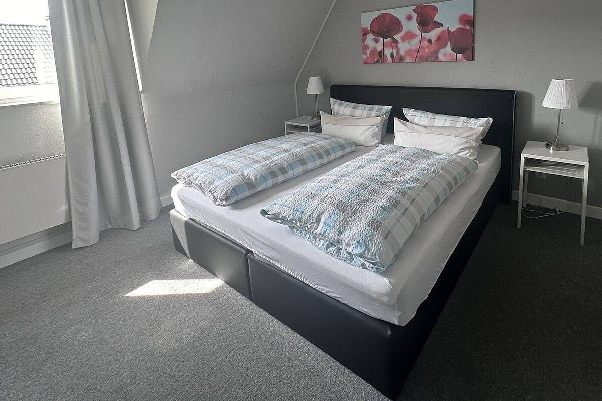 A neatly made bed with a checkered duvet, two bedside tables with lamps, a painting on the wall, and a window with curtains.