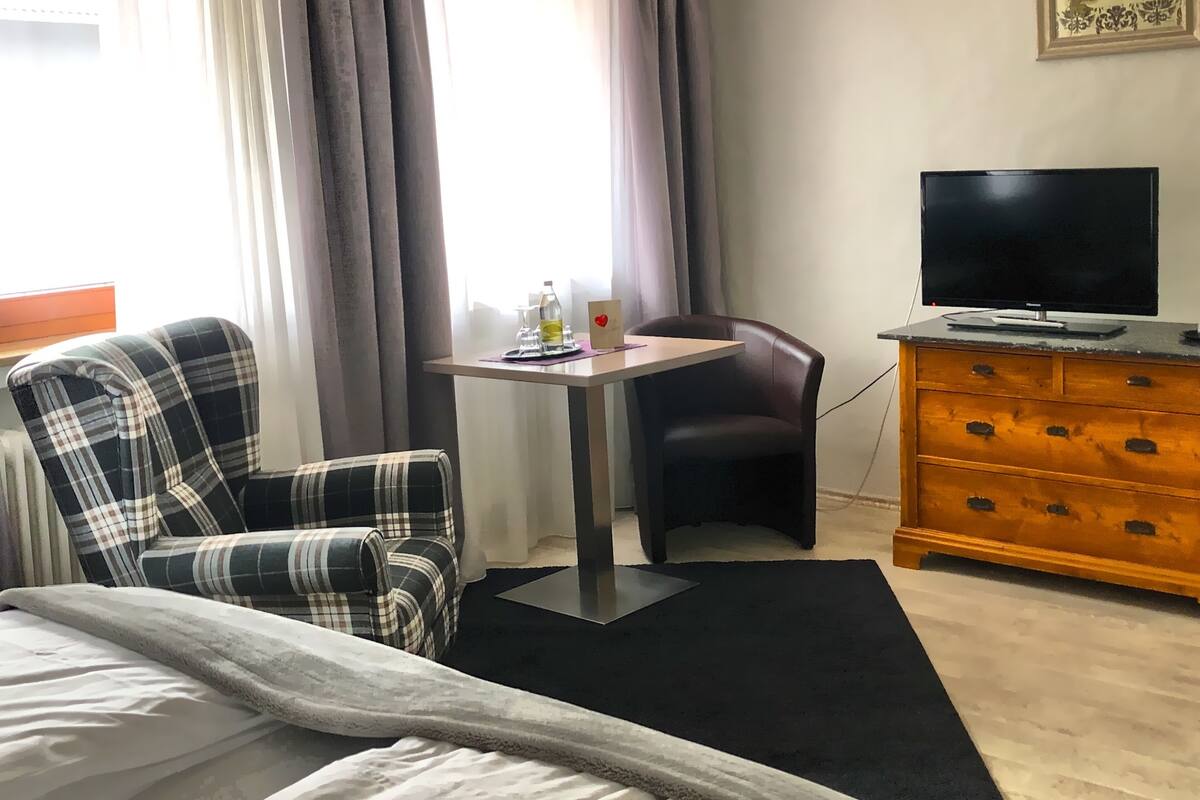 A hotel room with a bed, a plaid armchair, a small table with a bottle and glasses, a TV on a wooden dresser, and a window with curtains.