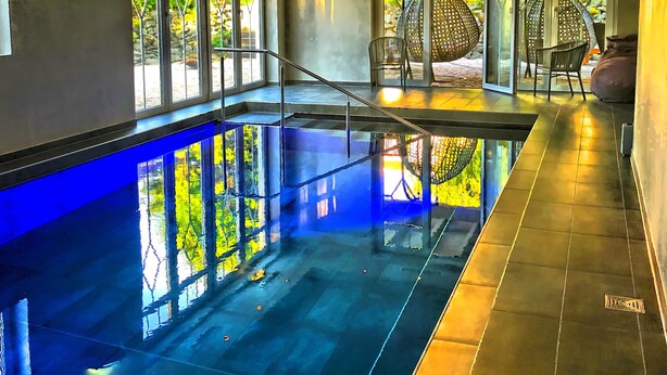 Una piscina con fondo de cristal, rodeada de paredes con ventanas amplias, y una silla y un sillón de plumas en la zona contigua.