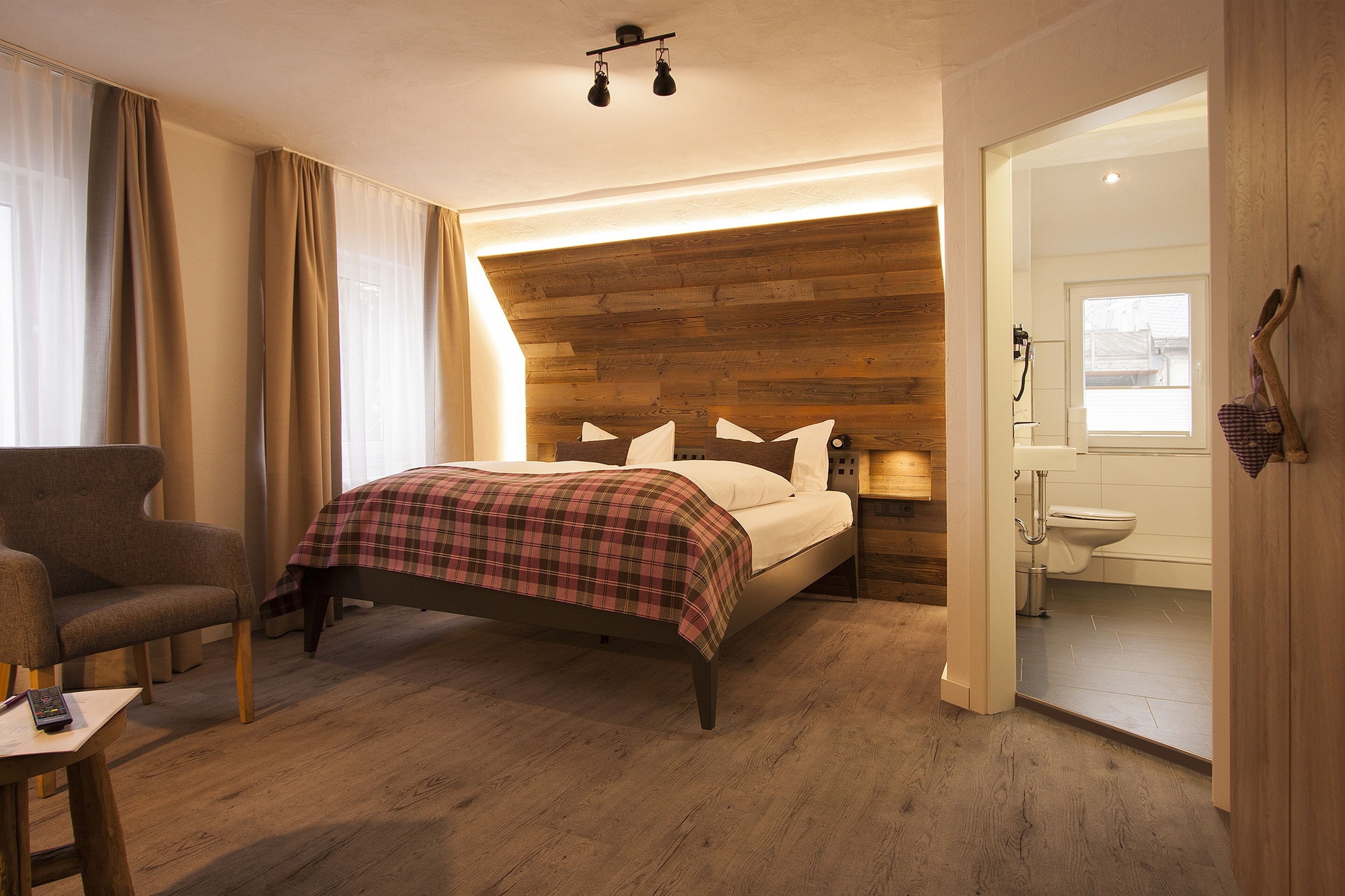 Un dormitorio con cabecera de madera, cama con manta escocesa, una silla y una mesita de noche con control remoto.