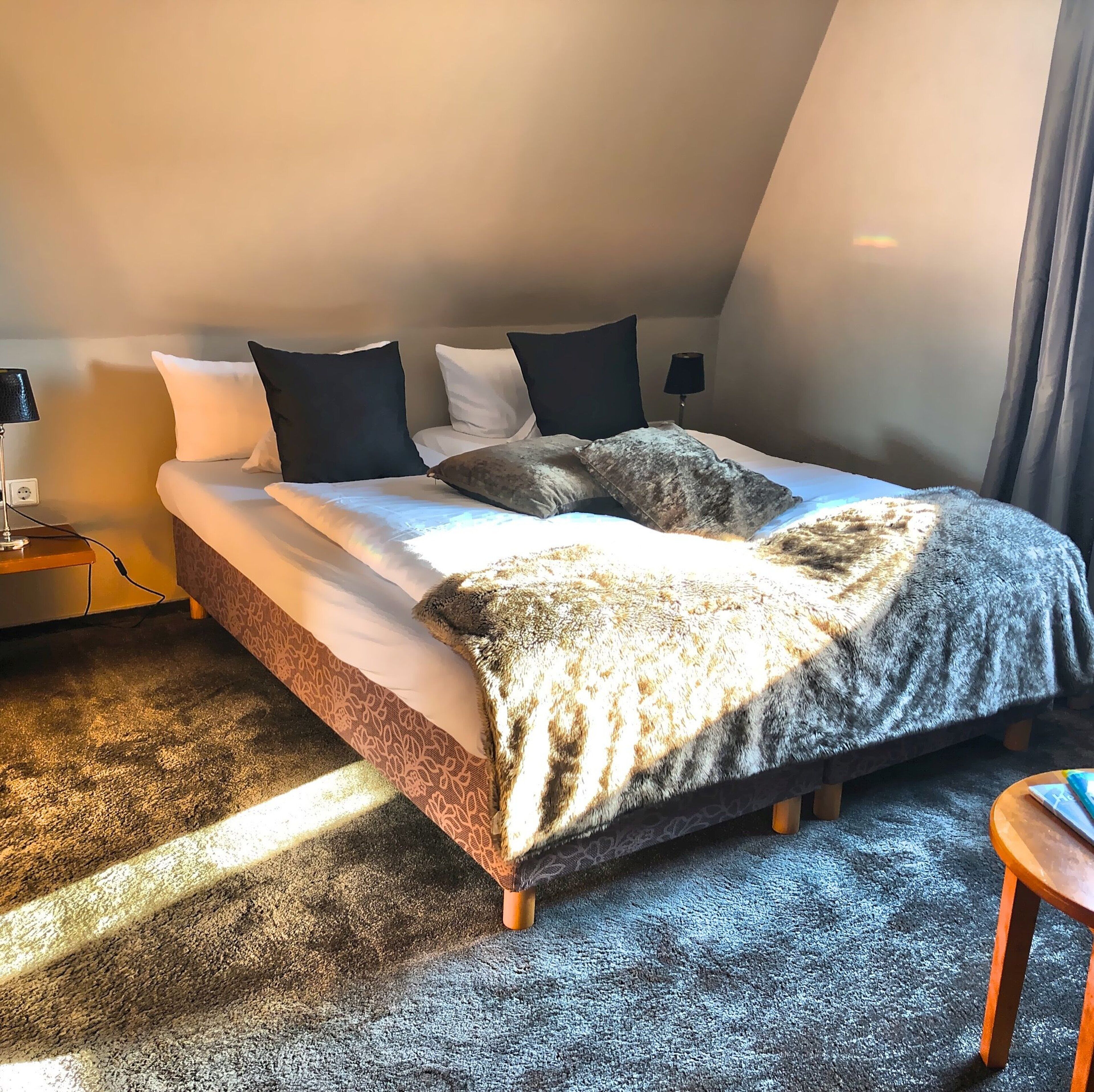 A neatly made bed with a patterned bedspread and multiple pillows, a bedside lamp, and a small wooden nightstand.