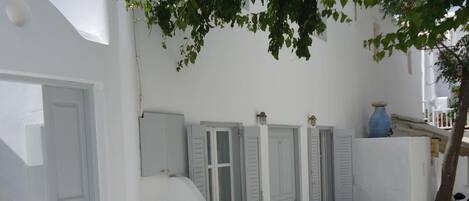 A white building with a stone floor, white doors, and shutters, and a tree providing shade.