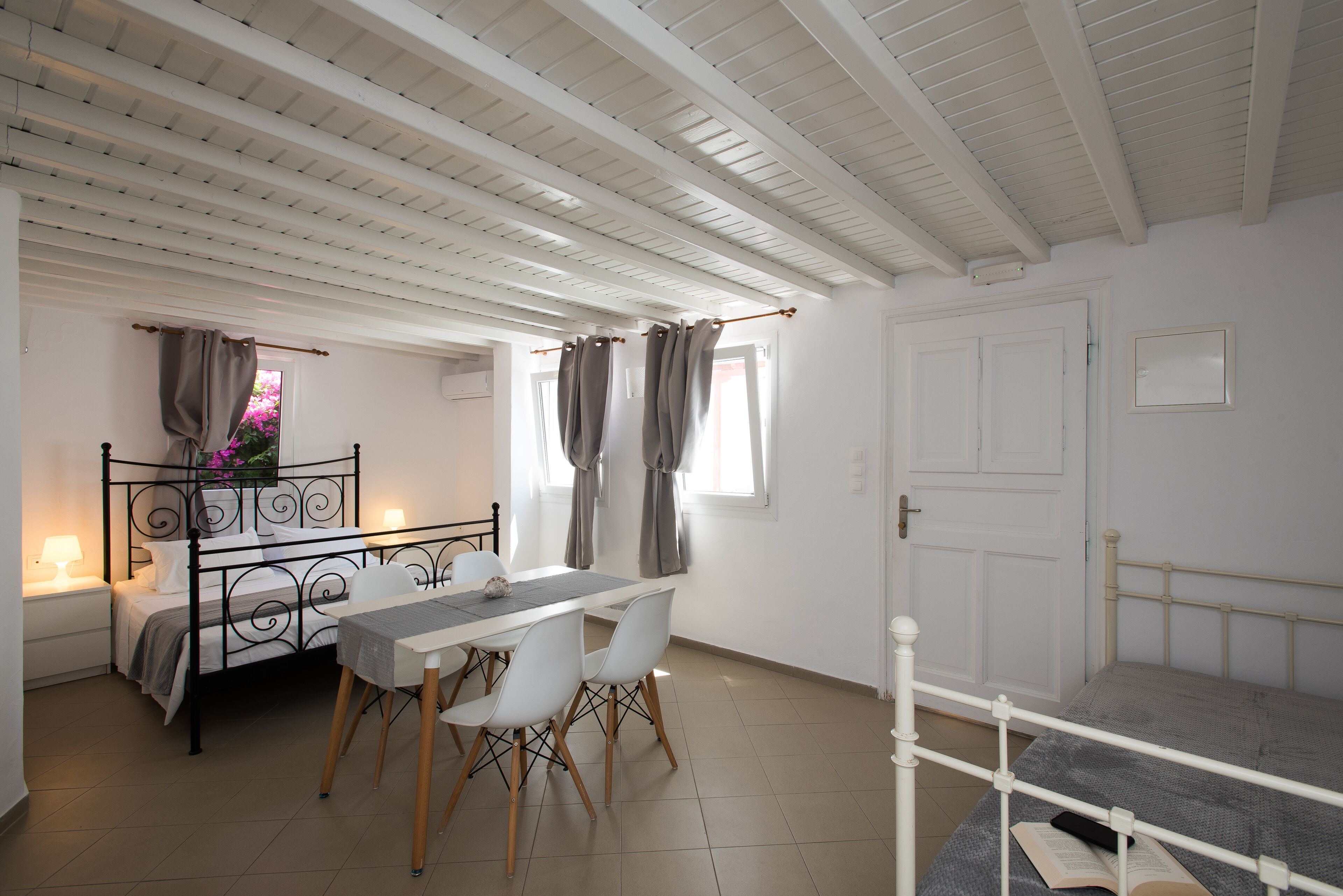 A bedroom with a bed, a bedside table, a desk with chairs, and a window with curtains.