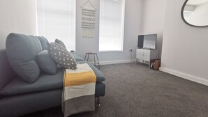 En moderne stue med en grå sofa, en lille skammel, et fjernsyn på et stativ og et spejl monteret på væggen.