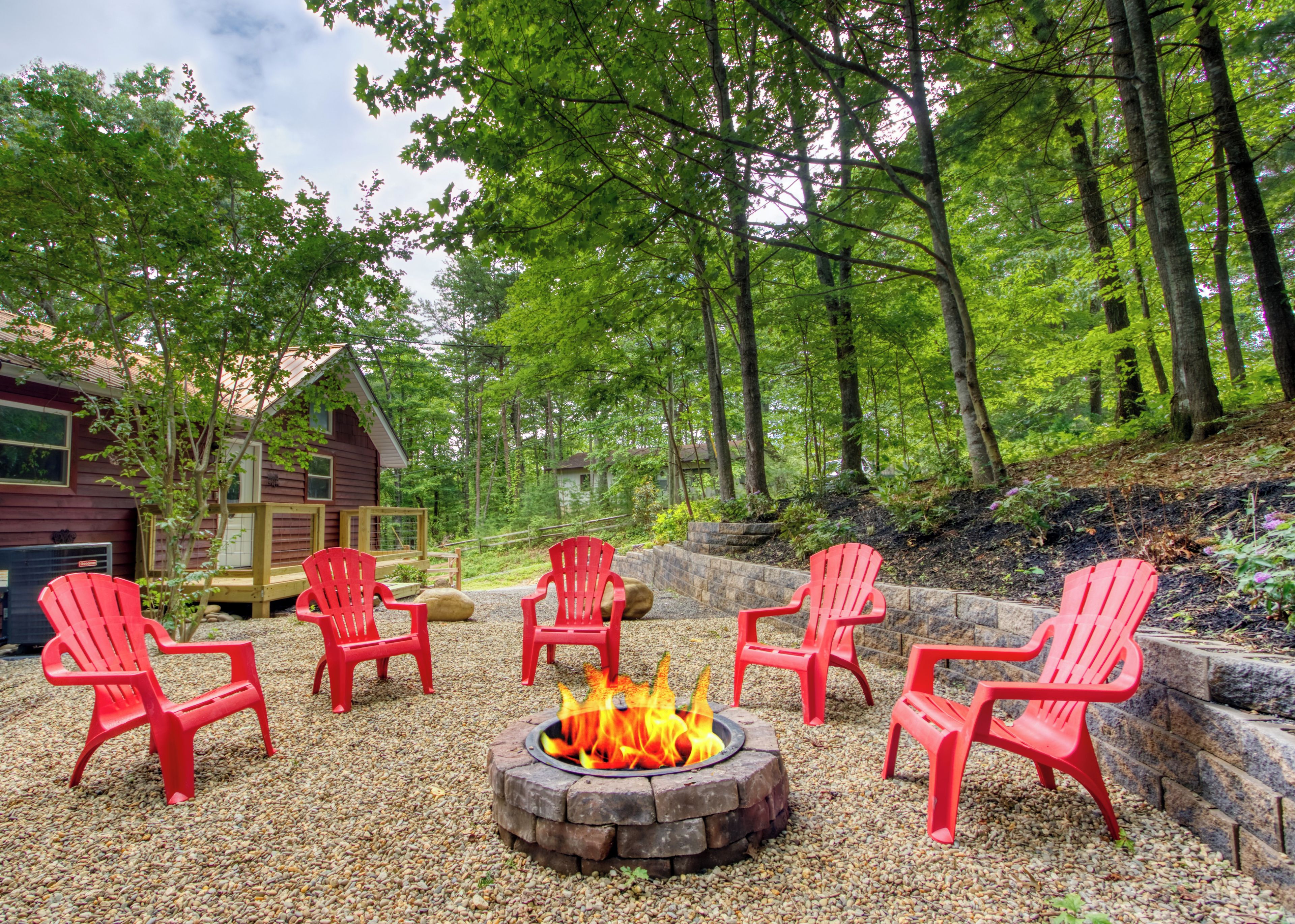 A forested outdoor area with a fire pit and red Adirondack chairs arranged around it.
