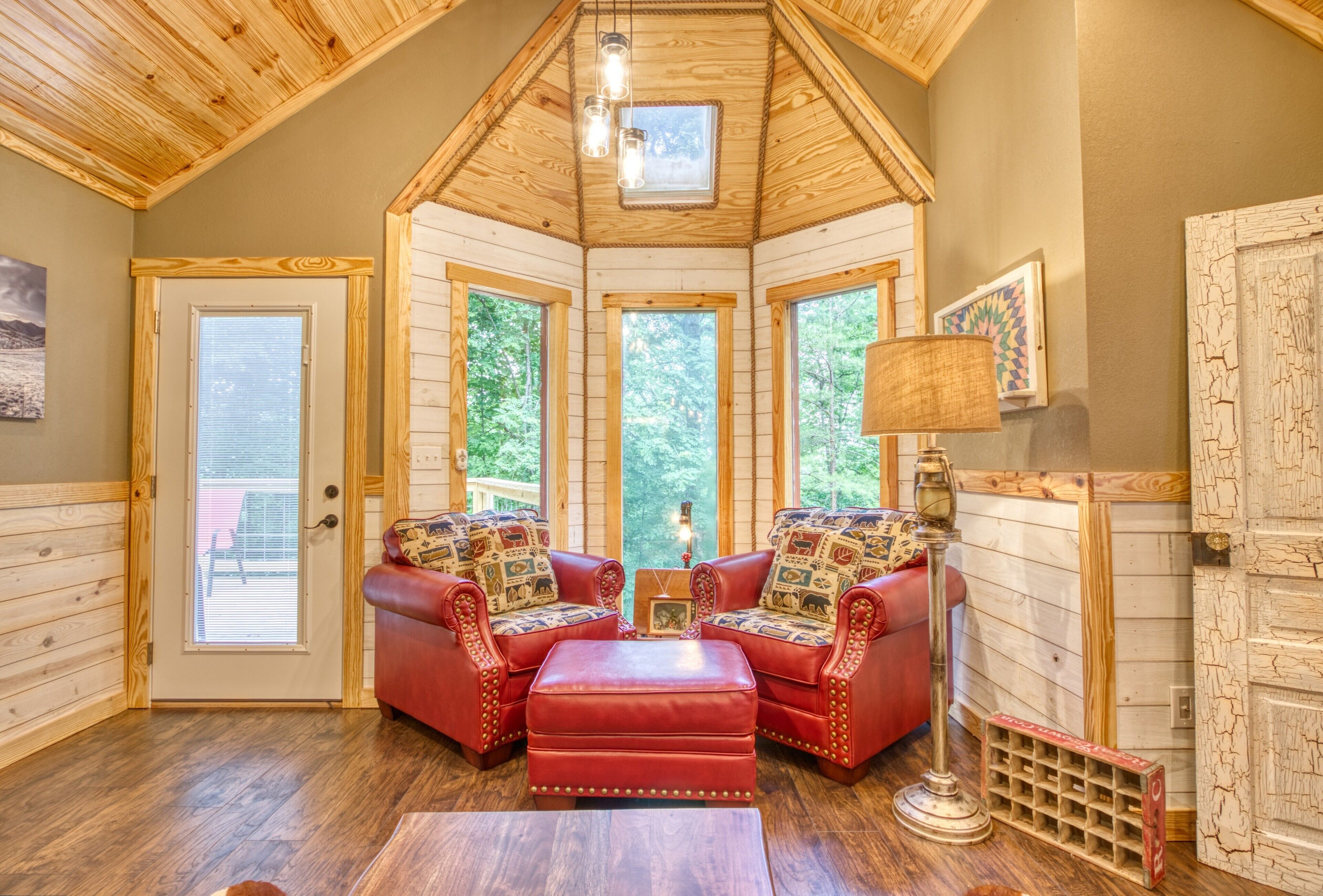 A cozy cabin interior with a wooden ceiling, large windows, a red leather sofa set, and a small table.