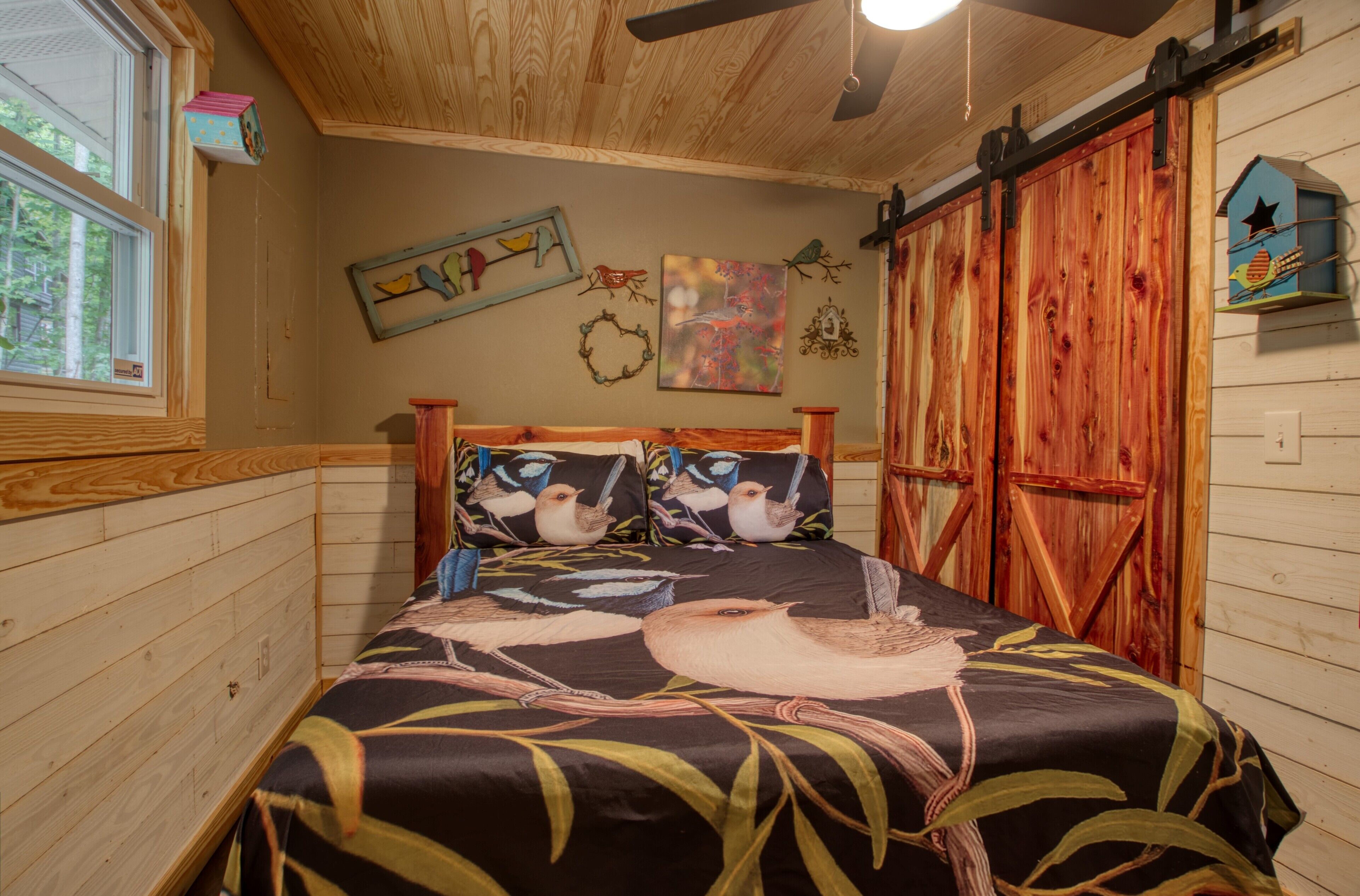 A bedroom with a bed featuring a bird-themed bedspread, wooden walls, a window, and a ceiling fan.