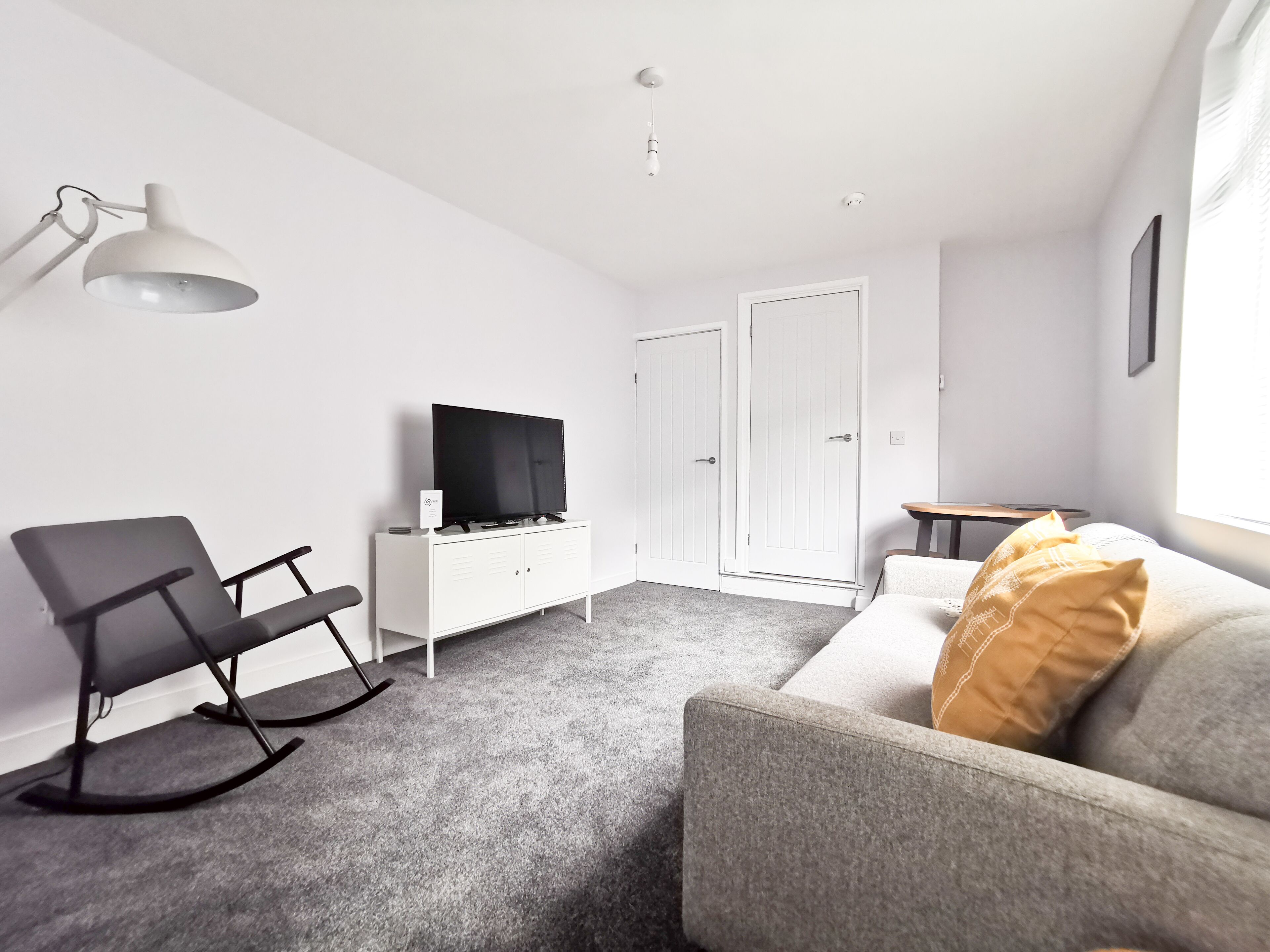 A modern living room with a grey sofa, a black rocking chair, a white entertainment unit with a TV, and a white wardrobe.