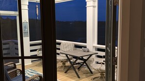 Una puerta corrediza de vidrio abierta que da a un patio exterior con una mesa y sillas, una silla de mimbre y una alfombra decorativa.