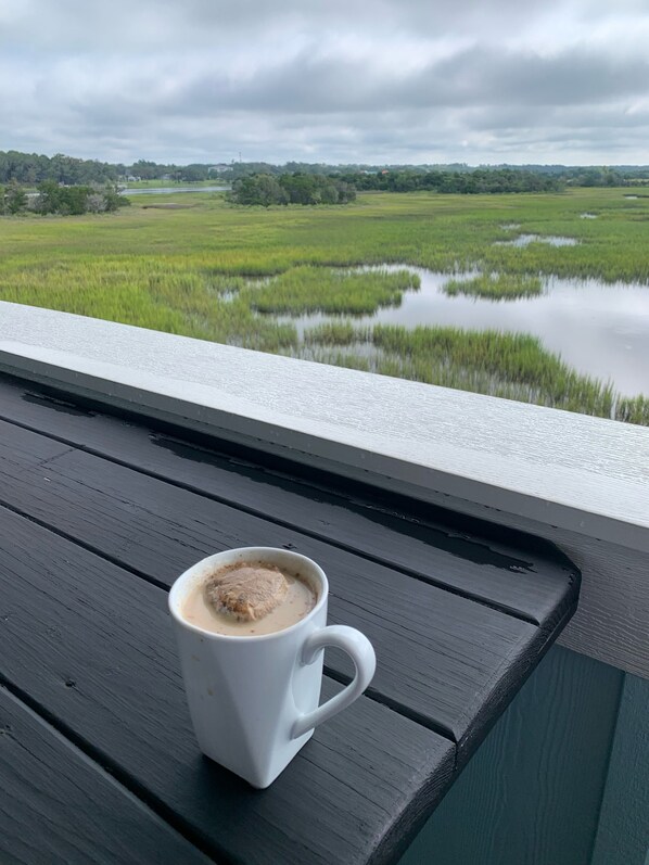 A white mug with a beverage on a wooden table, overlooking a green landscape with water bodies and trees. - Amazing marsh view and close to beach! (Holden Beach)