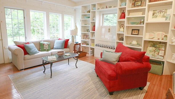 A living room with a striped sofa, a red armchair, and a coffee table. There are large windows and a bookshelf filled with various items.
