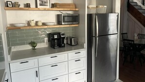 A modern kitchen with white cabinetry, a stainless steel refrigerator, a microwave, a coffee maker, and a toaster. There are shelves above the counter holding bowls and a framed picture. - Sweet Mary 2 Bedroom Home by RedAwning (Jamestown)