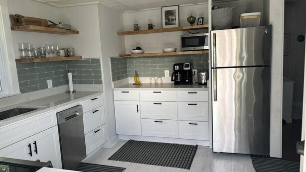 A modern kitchen with white cabinets, a stainless steel refrigerator, and a microwave. There are wooden shelves above the counter and a black and white striped rug on the floor. - Sweet Mary 2 Bedroom Home by RedAwning (Jamestown)