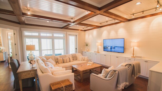 A spacious living area with a wooden ceiling, white walls, and a large window. There is a sofa set, a coffee table, and a TV mounted on the wall.