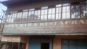 A building with a sign for 'Apex Bar & Restaurant' offering self-contained rooms, a hot shower, secure parking, and DSTV. - Apex Guest Rooms (Juja)