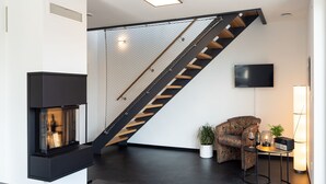 Una sala de estar moderna con chimenea, una escalera con acabado de madera, un televisor montado en la pared y una zona de estar con un sofá estampado y una mesita con una vela y una botella.