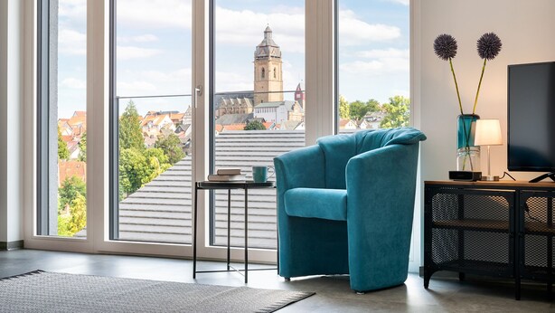 Una sala de estar moderna con un sillón verde azulado, una pequeña mesa con un libro y una taza, un televisor sobre un soporte negro y vistas de un paisaje urbano a través de amplias ventanas.