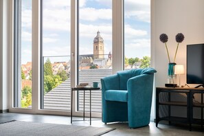 A modern living room with a teal armchair, a small table with a book and a mug, a TV on a black stand, and a view of a cityscape through large windows.