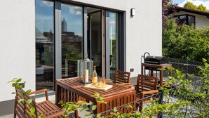 Una casa moderna con un patio que cuenta con una mesa y sillas de madera, una parrilla y vistas de la ciudad a través de amplias ventanas.