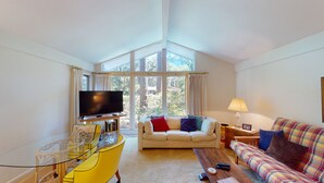 Una sala de estar con un ventanal grande, un televisor de pantalla plana, un sillón amarillo, un sofá y una lámpara.