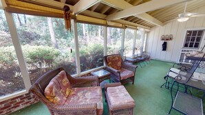 Un espacio de estar al aire libre con techo, con mobiliario de mimbre y una mesa, con vistas a una zona boscosa.