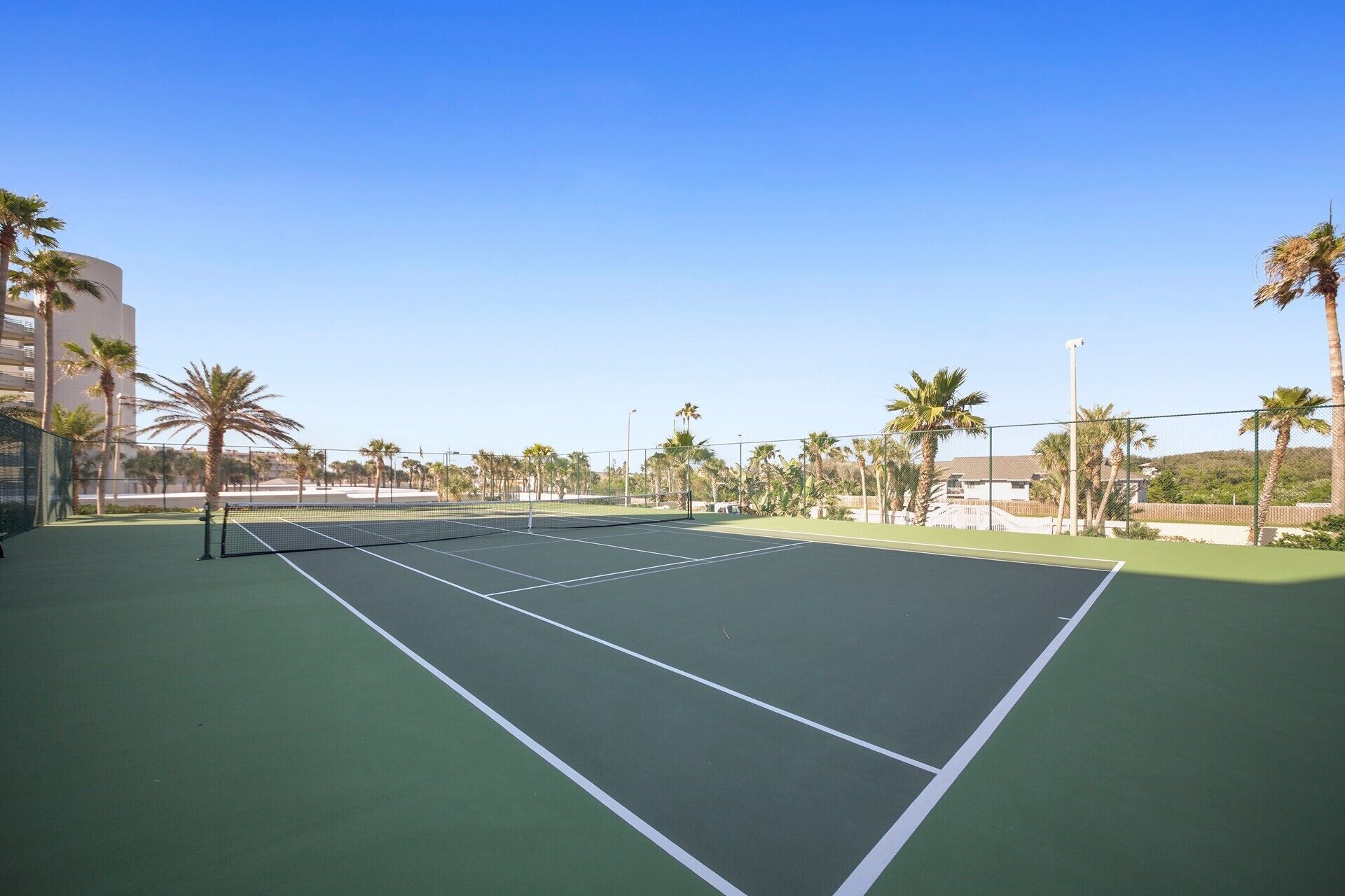A tennis court with a net, surrounded by palm trees and buildings.