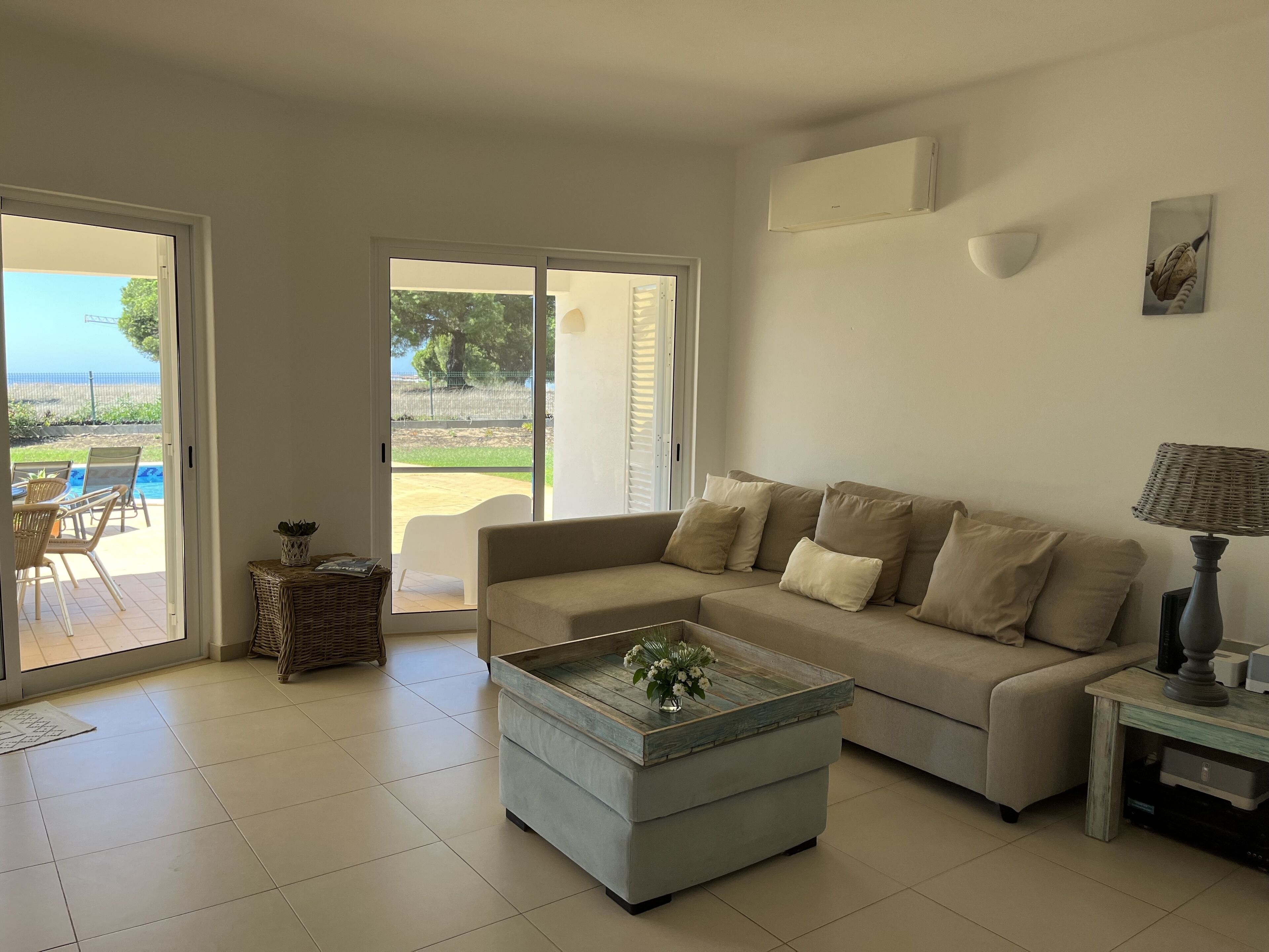 A living room with a beige sofa, a coffee table, and a wicker side table. There is a sliding glass door leading to an outdoor area with a pool and chairs.