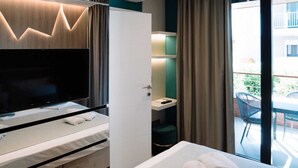 A modern hotel room with a flat-screen TV, a bed with a white mattress and pillows, a small bedside table, a built-in bench, a large window with curtains, and a view of the outdoor area. - Lena Luxury Apartments (Zadar)