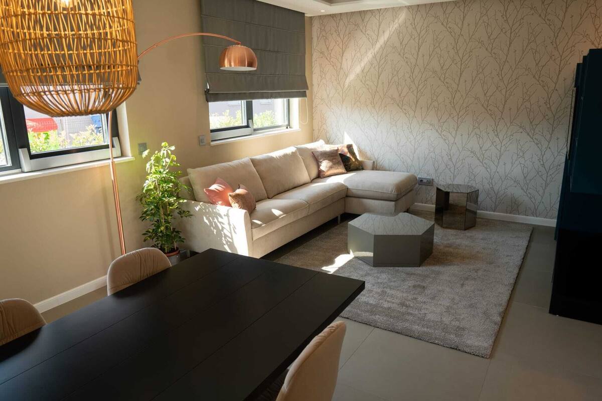 A modern living room with a sofa, a dining table, and a coffee table. There is a large window providing natural light and a view of the outdoors.