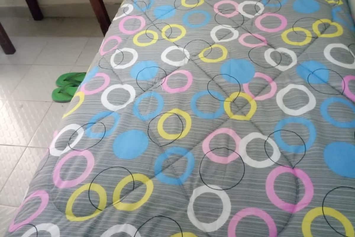 A small, single-bed room with a grey bedspread featuring colorful circular patterns. A wooden nightstand with a remote control and a cup is visible.