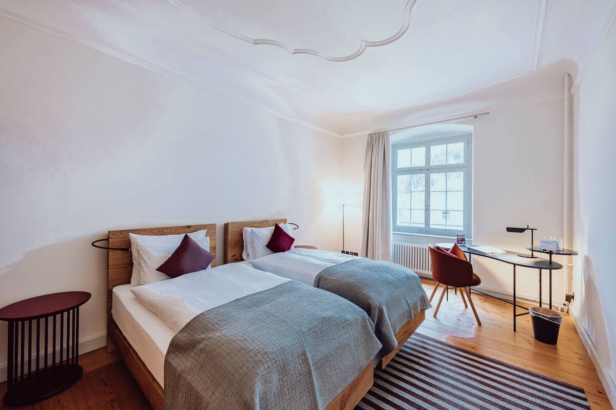 A hotel room with two beds, a desk, a chair, and a large window.
