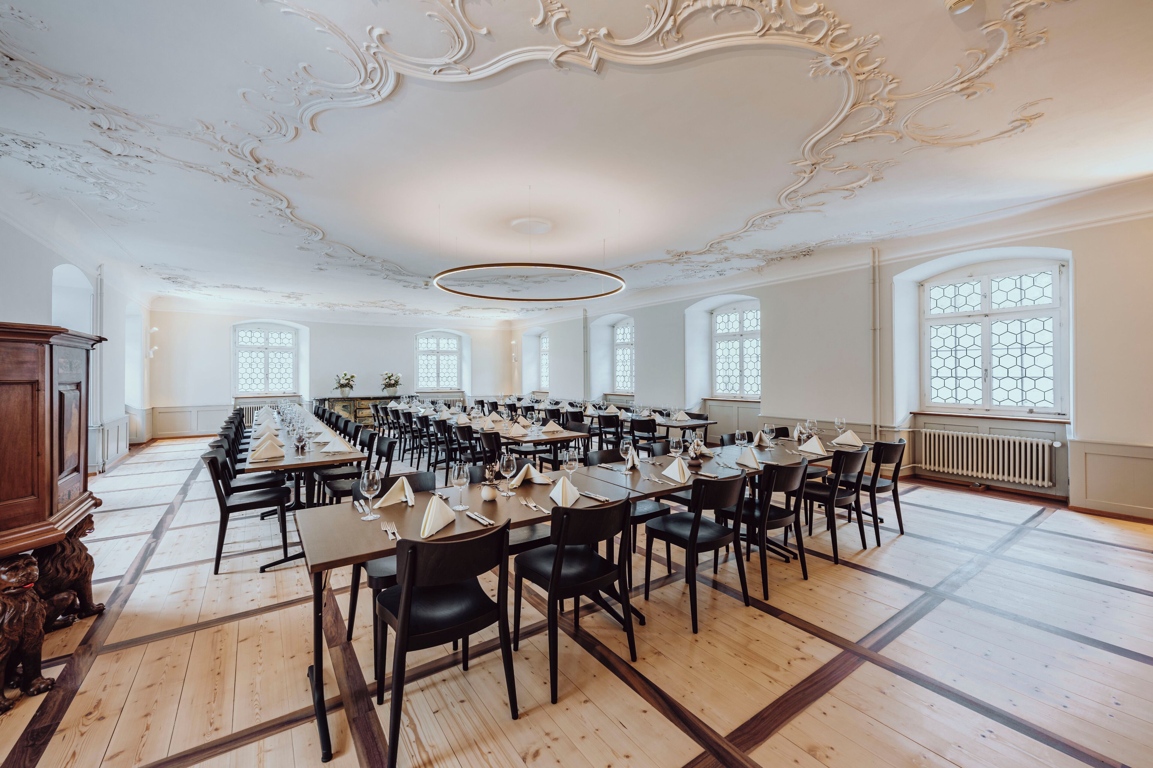 A large dining hall with long tables, black chairs, and ornate ceiling details.