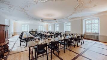A large dining hall with long tables, black chairs, and ornate ceiling details.