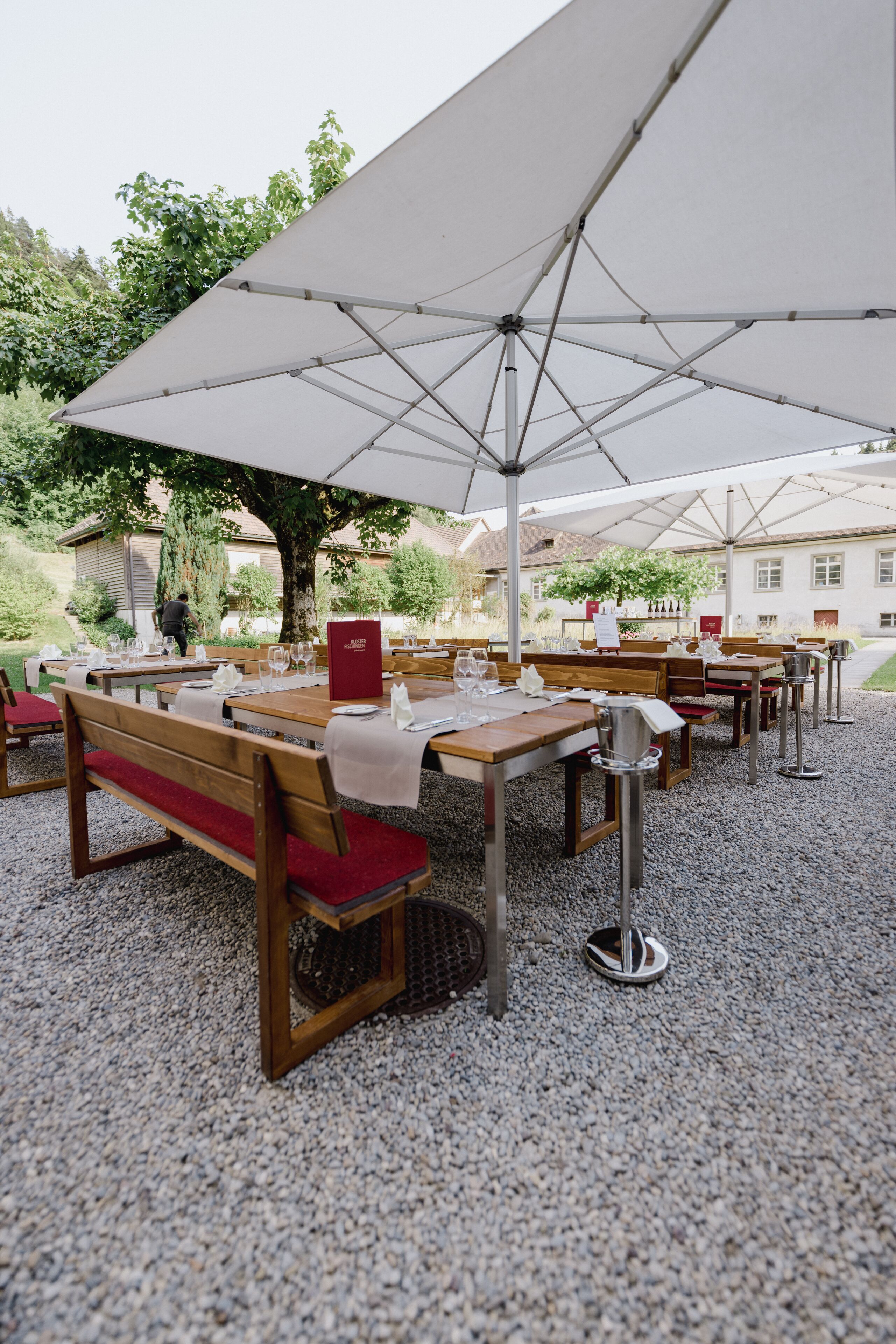 An outdoor dining area with tables and benches, covered by large umbrellas, set on a gravel surface.