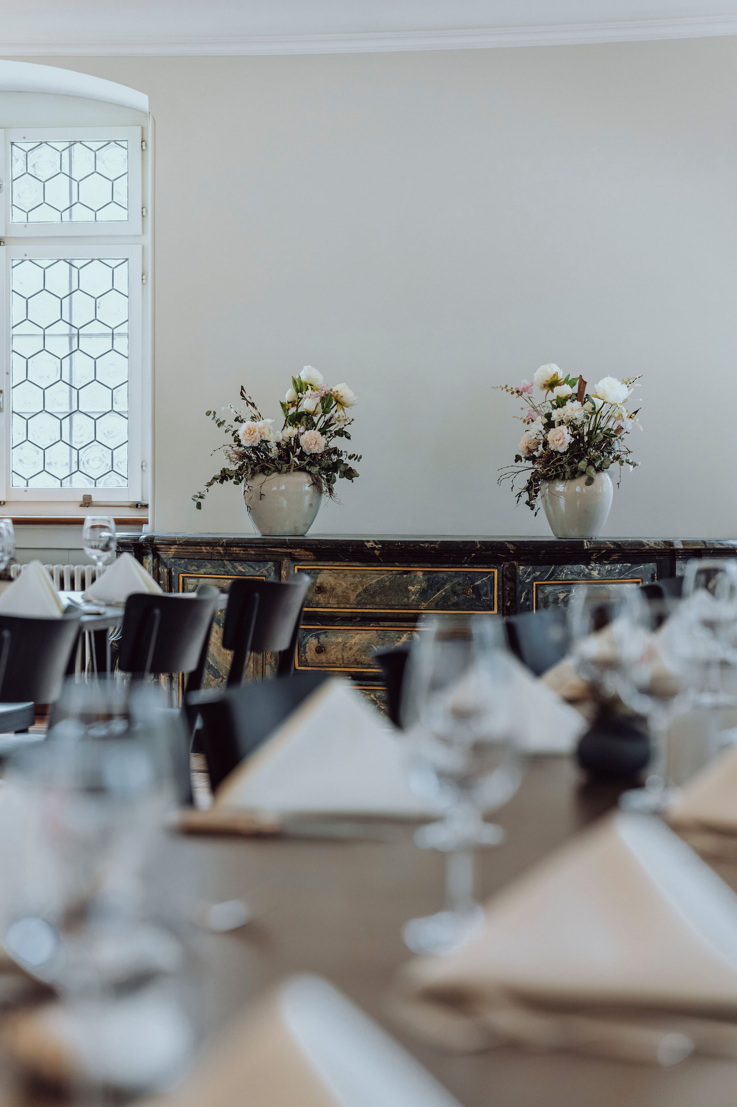 Een eetruimte met een lange tafel gedekt voor een maaltijd, twee vazen met bloemen en een raam met een honingraatpatroon.