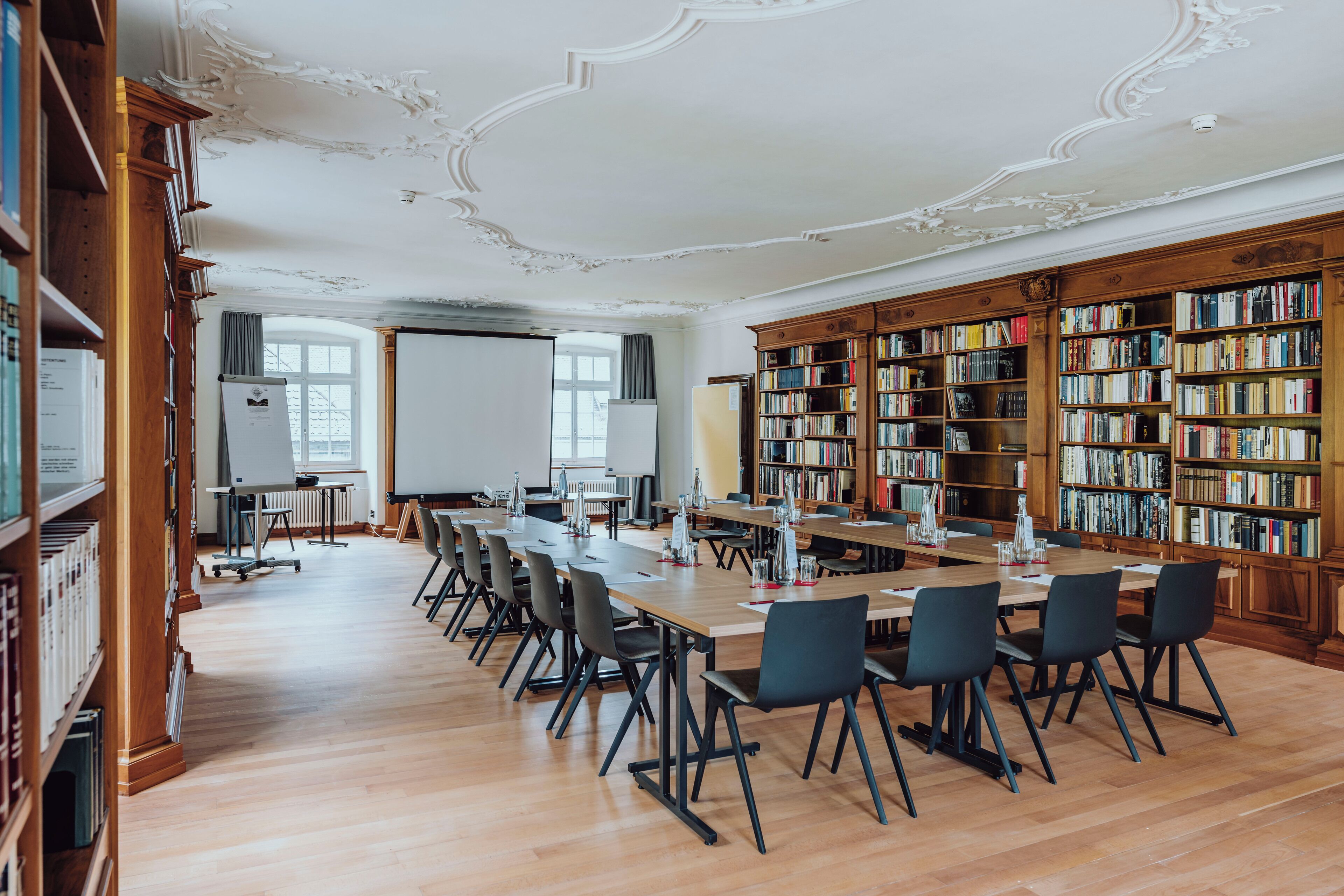 A library with wooden bookshelves, a long table with chairs, and a whiteboard.