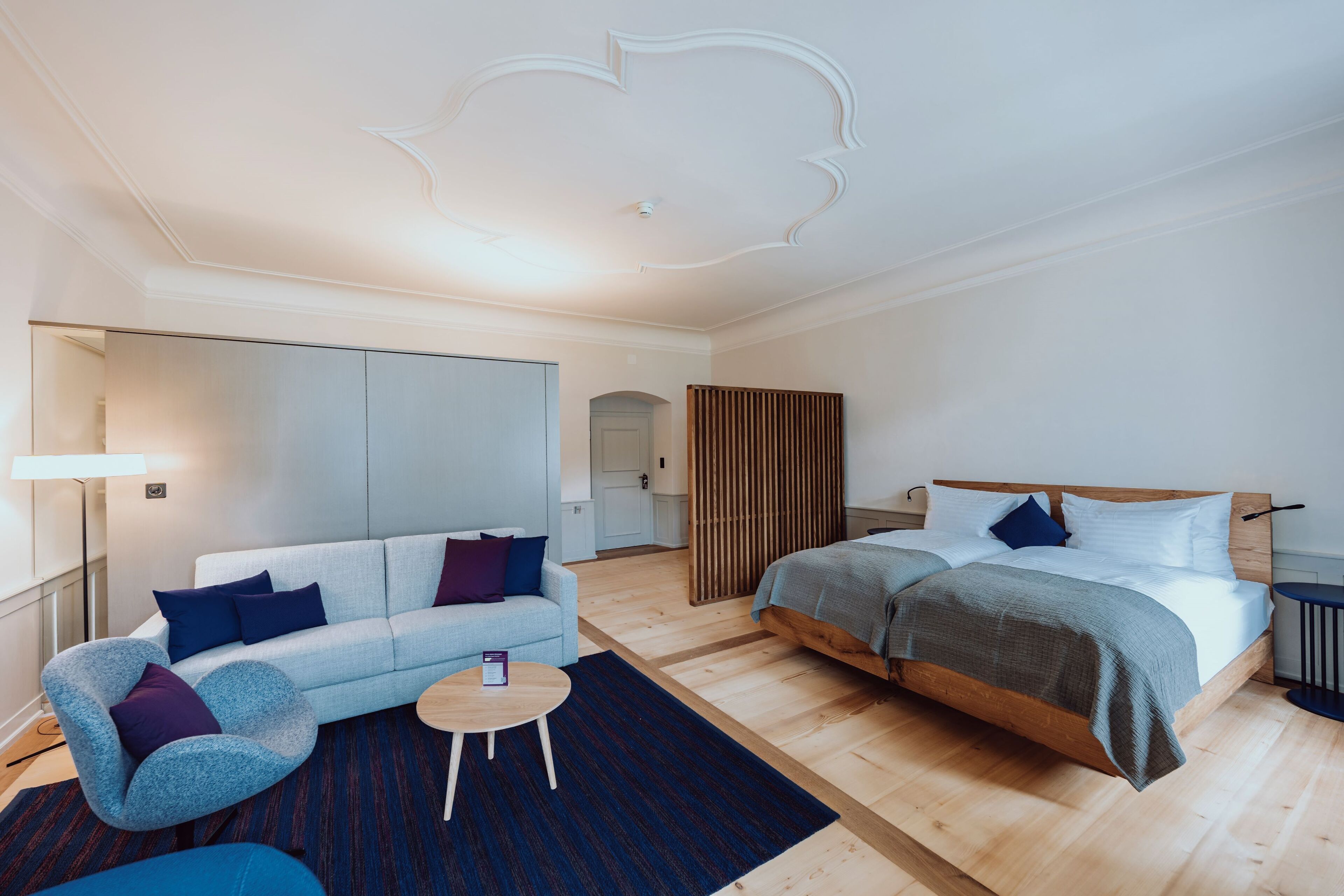 A modern hotel room with a sofa, a round coffee table, a bed with a wooden headboard, and a large mirror.