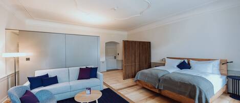 A modern hotel room with a sofa, a round coffee table, a bed with a wooden headboard, and a large mirror.
