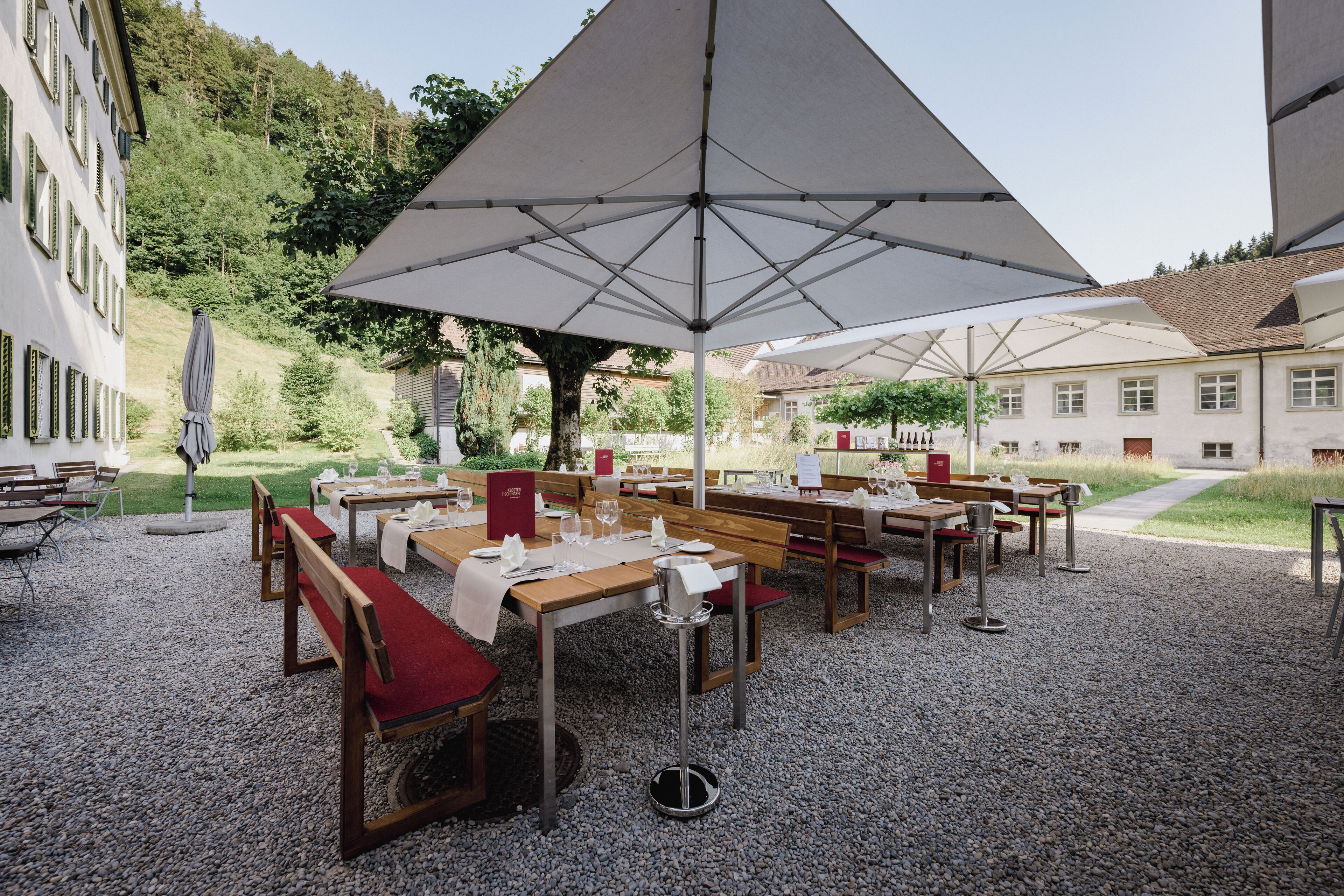 An outdoor dining area with tables and benches, surrounded by a gravel path and buildings.