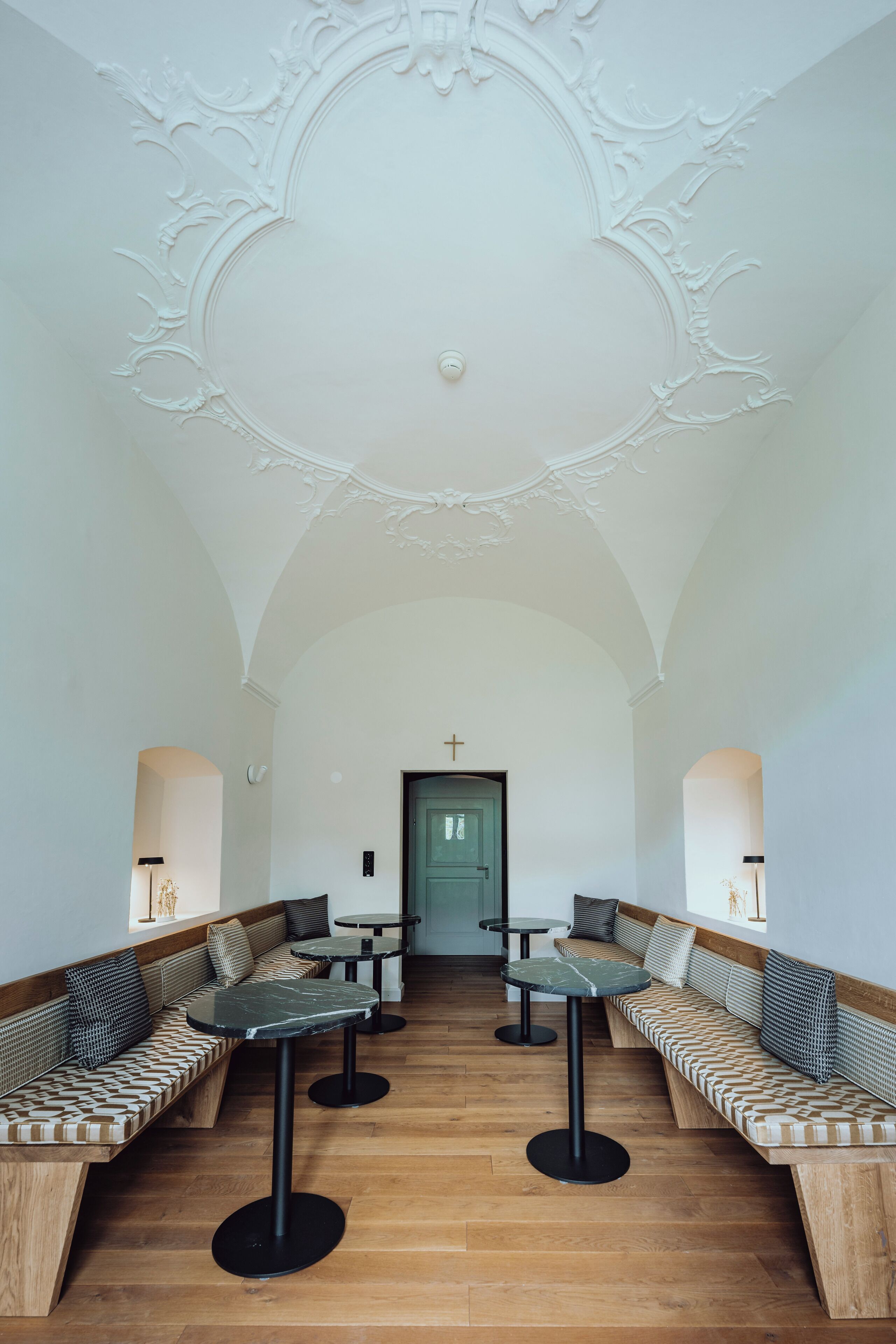 A spacious interior with high ceilings, ornate ceiling details, and a central green door. There are multiple seating areas with patterned cushions and round tables.