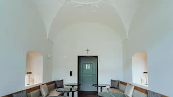 A spacious interior with high ceilings, ornate ceiling details, and a central green door. There are multiple seating areas with patterned cushions and round tables.