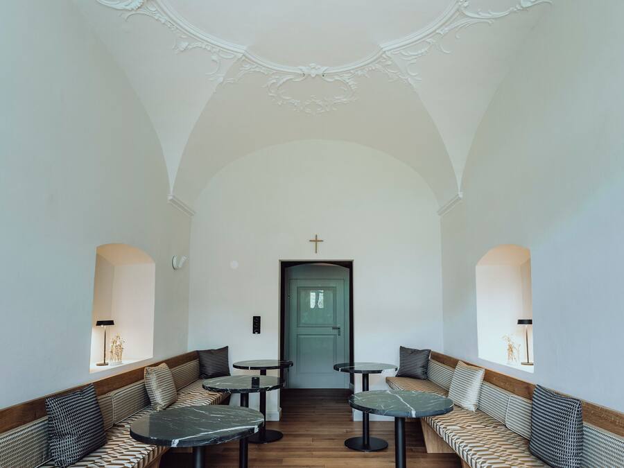 A spacious interior with high ceilings, ornate ceiling details, and a central green door. There are multiple seating areas with patterned cushions and round tables.