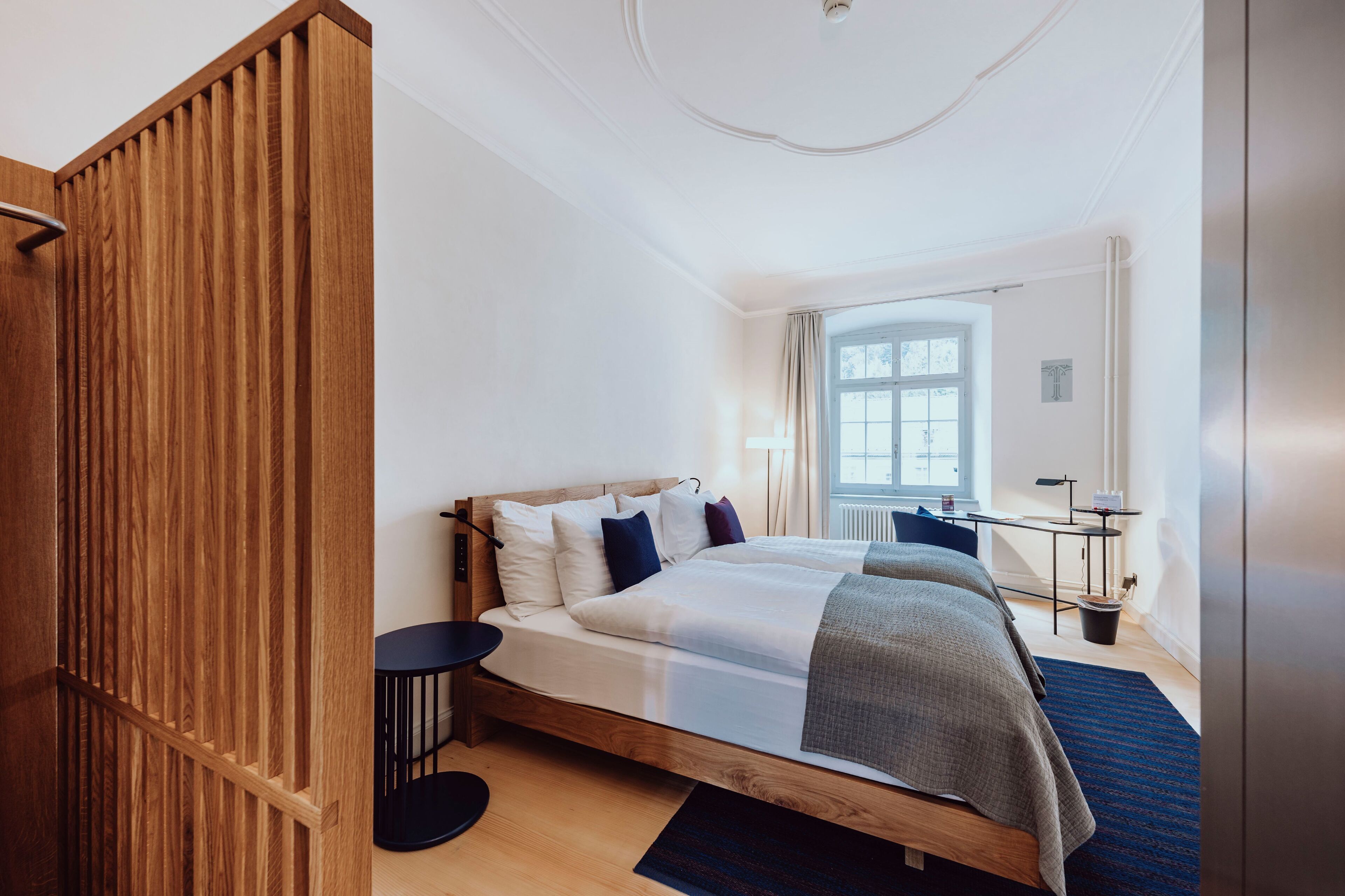 Quarto de hotel moderno com uma cama de madeira, uma secretária pequena e uma janela grande.